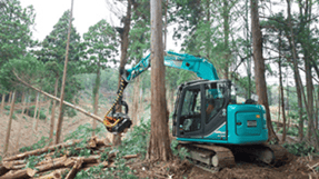 山林の中で、木材をつかんで作業を行う林業用油圧ショベルの実機写真。