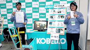 「くるくるCOOL君」のパネルがあるブースで、2名のスタッフが賞状と目録を掲げている写真。