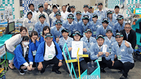 展示会場での集合写真。法被を着たスタッフや作業服のメンバーが賞状を囲んでいる様子。