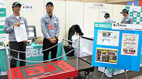 展示ブースで「持ってRUN」のパネル横に、グレーの作業服を着た男性2名が立っている写真。