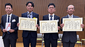 表彰状などを手にし、並ぶスタッフ4名の集合写真。