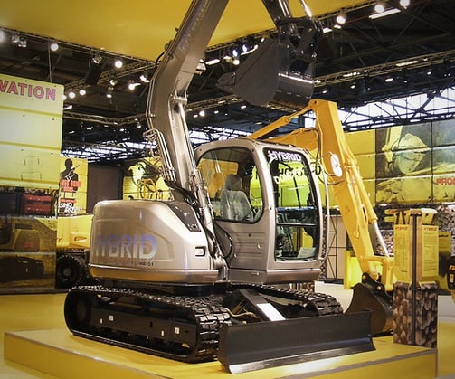 世界初のハイブリッドショベルモニター機「SK70H」または量産機「SK80H」。キャビン付近に『HYBRID』のロゴが入ったシルバーと黄色のショベルカーが、展示会場の台座の上に展示されている様子。