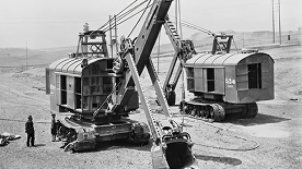 コベルコ建機の歴史を感じさせるモノクロ写真。広大な土木工事現場で稼働する、初期モデルの大型油圧ショベル2台と作業員の姿が写し出されており、長年にわたる建設機械開発の歩みと実績を象徴するイメージ画像です。